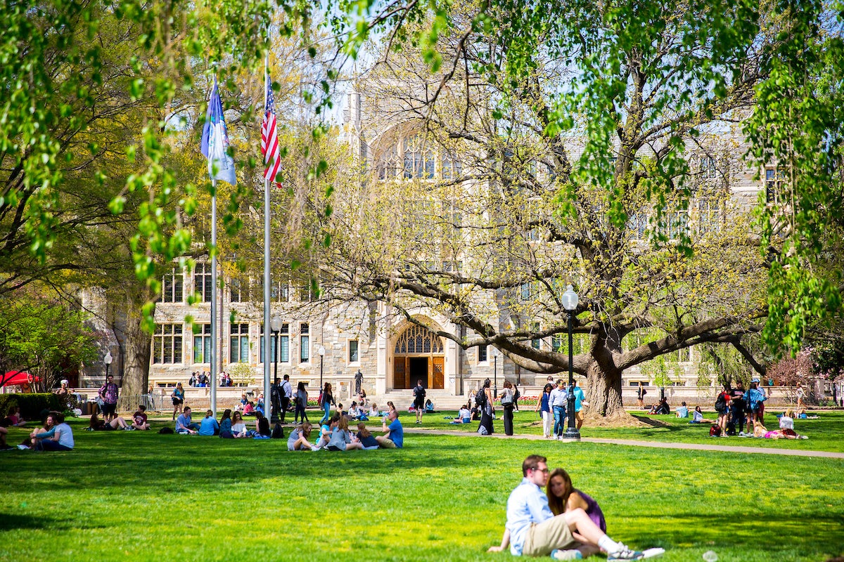 Students sitting on green lawn below green trees beside large stone, white building